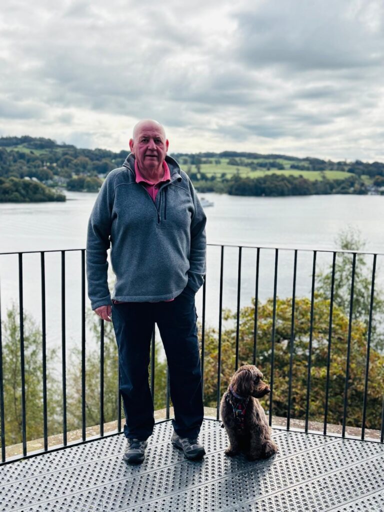 Robin Hebden standing on a decking in front of a lake with his pet dog sat by his side.