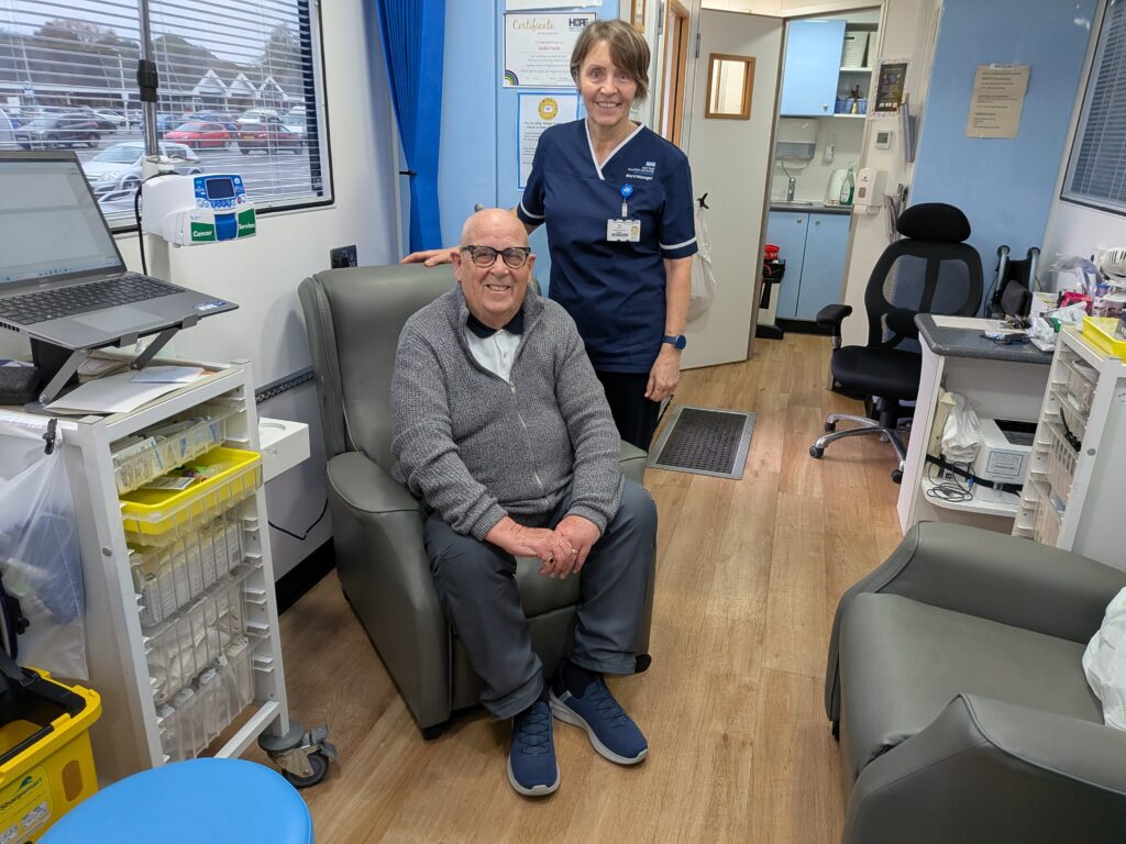Alan Morris sat in one of the grey treatment chairs on board his local mobile cancer care unit, arms in his lap and accompanied by one of the nurse team members standing behind the chair.