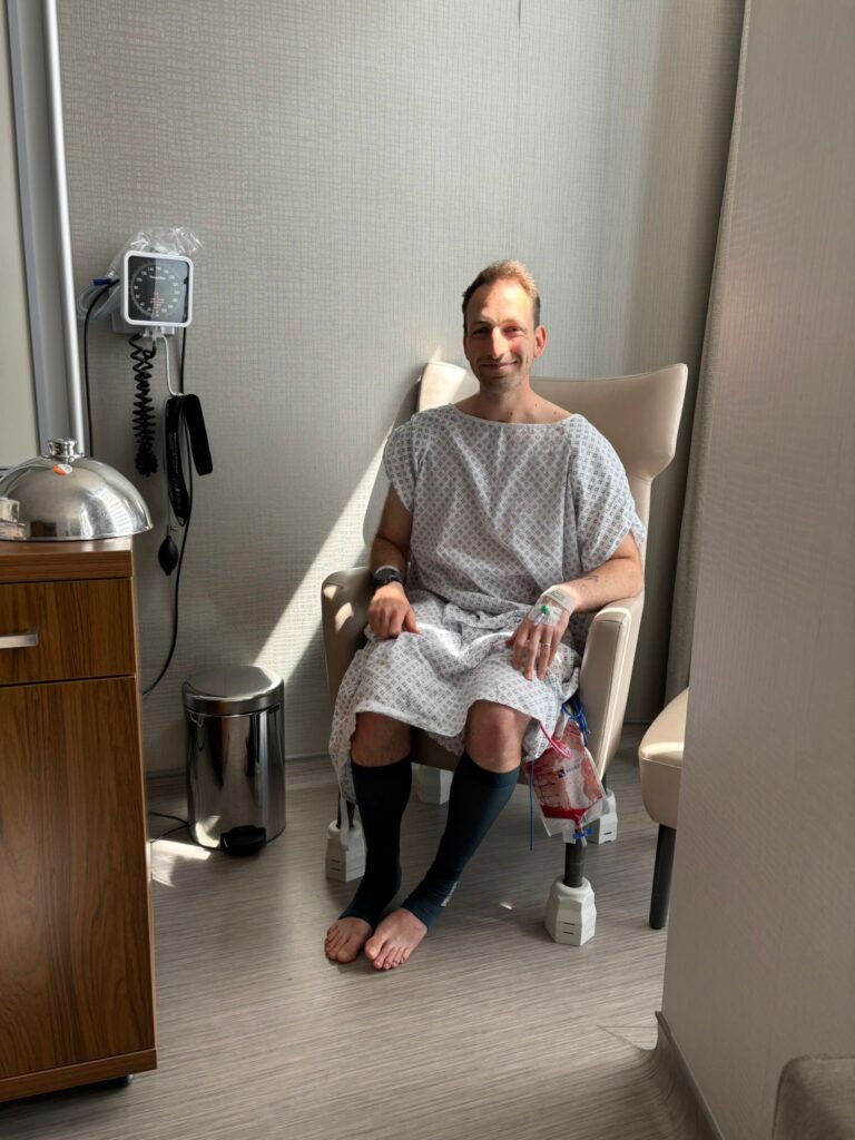 Luke sat in a hospital chair in a hospital gown after going through surgery