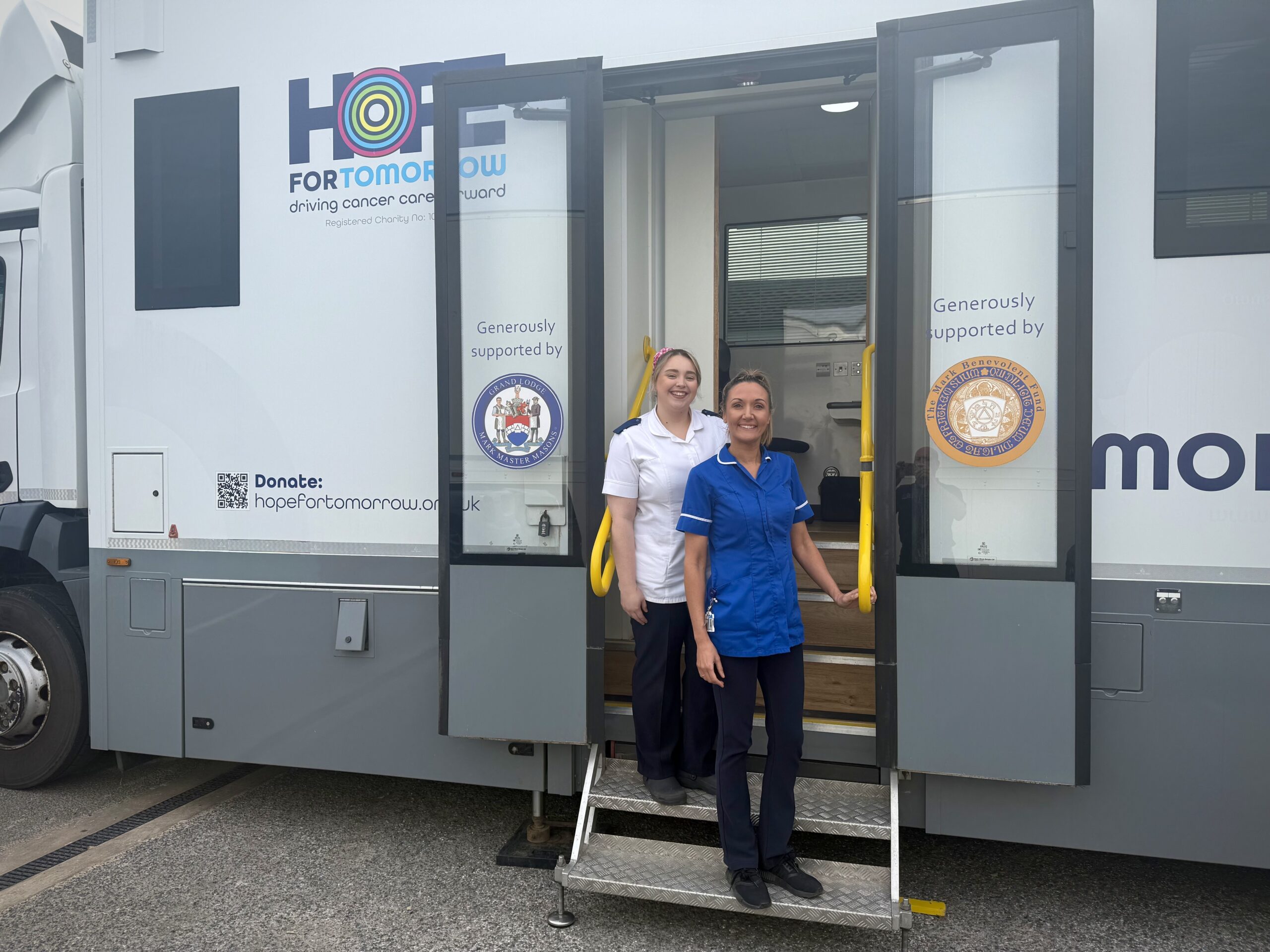 An image showing two NHS nurses smiling and standing side by side at the doorway to the mobile cancer care unit that they work on board. Anne-Marie Brooks is shown on the right. 